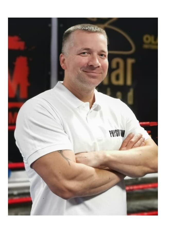 Smiling man with crossed arms wearing a white polo shirt, standing in a boxing gym.